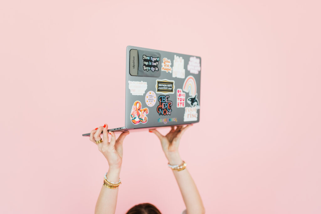 Flodesk Review: The Email Marketing Platform I Actually Use and Love 2 Hands raising a sticker-covered laptop against a pink background—symbolizing creative freedom in this Flodesk review.