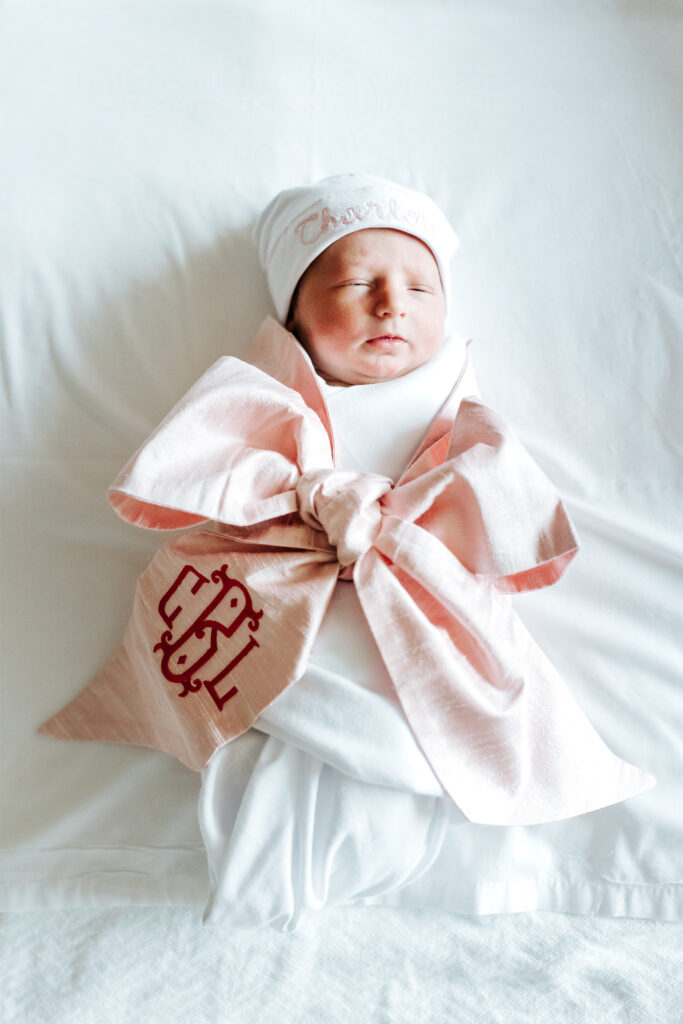 How to Create a Stress-Free Newborn Session at Home 1 A swaddled baby in a pink bow during an in-home newborn session, resting peacefully on a white bed.