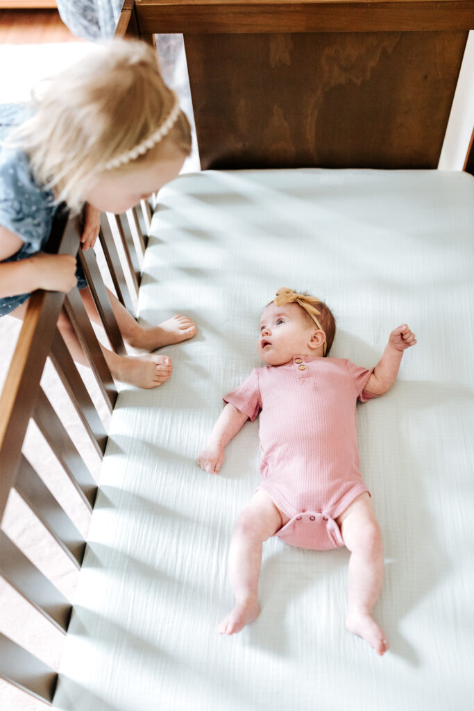 How to Create a Stress-Free Newborn Session at Home 5 Big sister peeking into the crib to look at her newborn sibling during a cozy family moment.