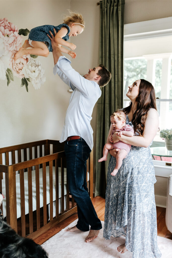 How to Create a Stress-Free Newborn Session at Home 6 Parents smiling as dad lifts toddler in the nursery while mom holds the baby—capturing playful energy during a newborn session.