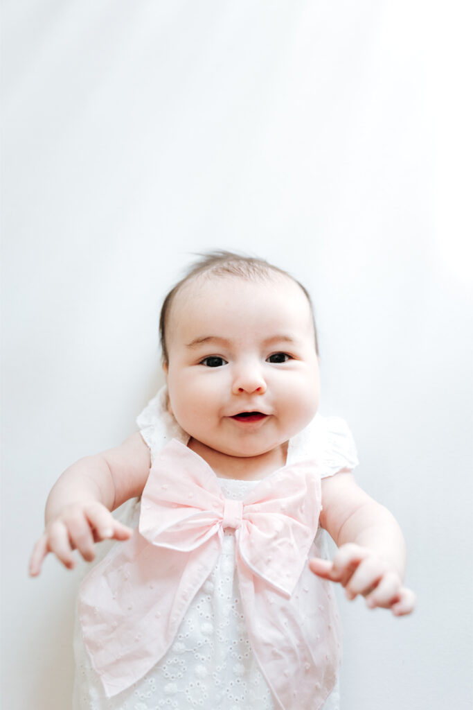 How to Create a Stress-Free Newborn Session at Home 3 Smiling baby wearing a white outfit with a pink bow, captured in bright natural light during a newborn session.