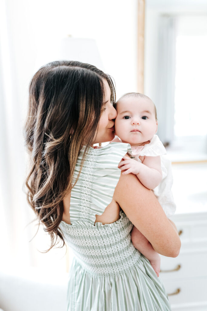 How to Create a Stress-Free Newborn Session at Home 4 Mother holding and kissing her baby’s head near a sunlit window, capturing a tender and timeless newborn session moment.